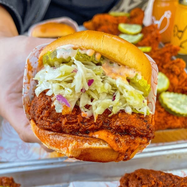 A hand holds a Cluck Kitchen fried chicken sandwich with coleslaw, pickles, and spicy sauce.