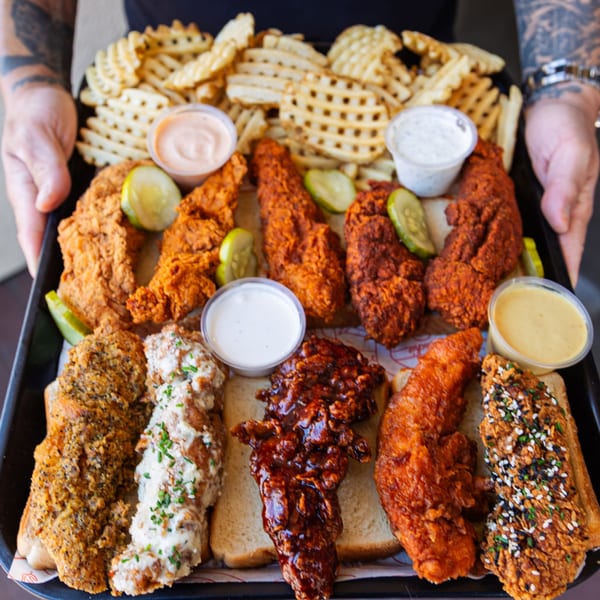 A large tray of Cluck Kitchen's fried chicken tenders with waffle fries, pickles, and dipping sauces.