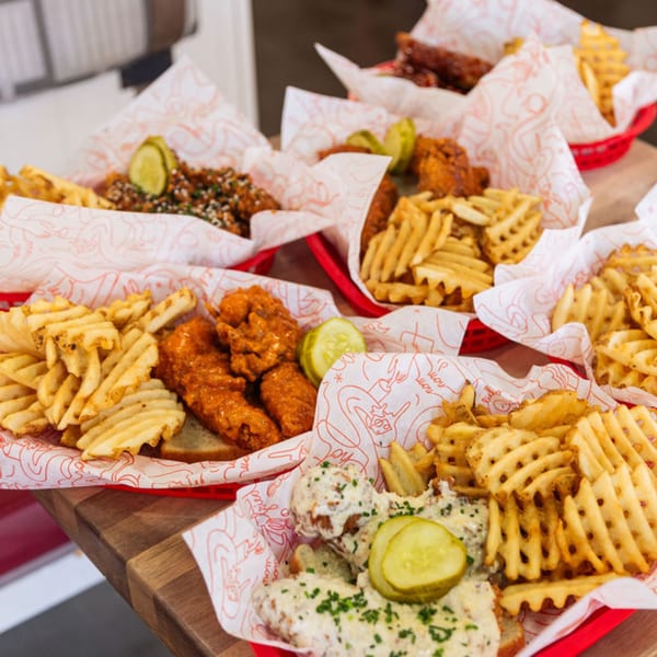 Multiple red baskets of Cluck Kitchen's fried chicken, waffle fries, and pickles on a wooden table.