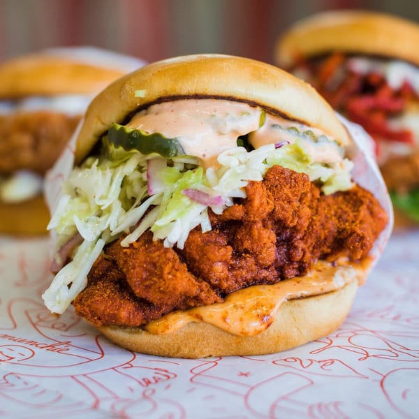 A Cluck Kitchen fried chicken sandwich with coleslaw, pickles, and sauce on a bun.