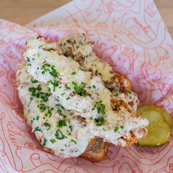 Cluck Kitchen fried chicken tenders covered in white sauce and chives, served with pickles on patterned paper.