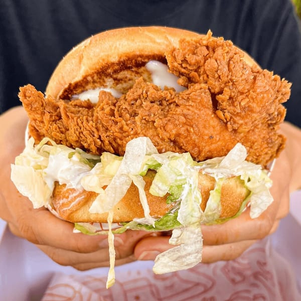 Hands holding a Cluck Kitchen fried chicken sandwich with lettuce and white sauce.