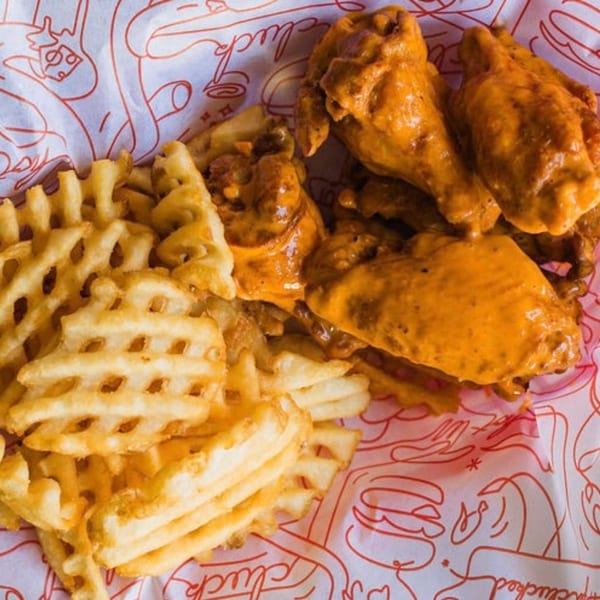 Cluck Kitchen's fried chicken wings coated in orange sauce, served with waffle fries on branded paper.